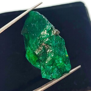 May include: A large, rough, green emerald gemstone. The emerald is held by tweezers against a black background.