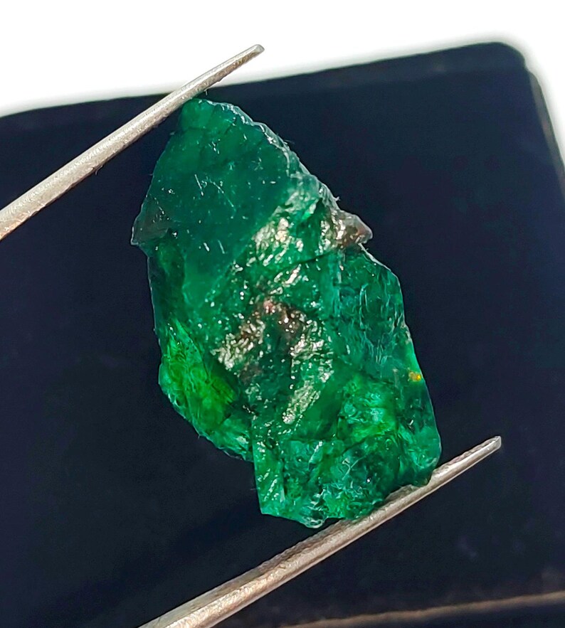 May include: A large, rough, green emerald gemstone. The emerald is held by tweezers against a black background.