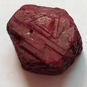 May include: A deep red ruby gemstone with a rough, faceted surface. The ruby has a natural, unpolished look and is a beautiful example of this precious stone.