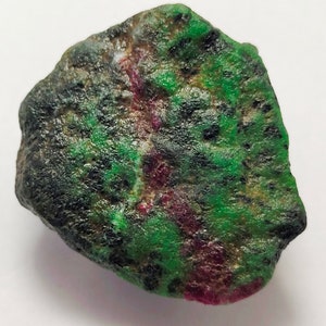 Op de afbeelding: Een ruwe, groene en roze edelsteen met een donkergroen en zwart gevlekt oppervlak.