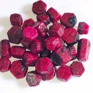 May include: A pile of raw ruby gemstones. The stones are a deep red colour, with some showing darker patches. The shapes vary, and they are scattered against a white background.
