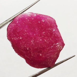 Può includere: Un grande rubino grezzo rosa con una forma leggermente irregolare. Il rubino è tenuto da una pinzetta su uno sfondo bianco.