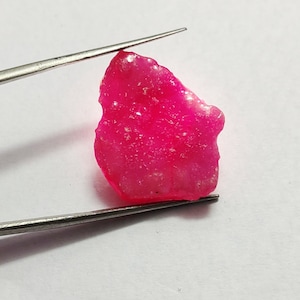 Puede incluir: Una gema rosa en bruto con un acabado brillante y translúcido. La gema está sujeta por unas pinzas sobre un fondo blanco.
