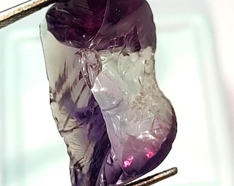 May include: A faceted purple and green gemstone with a rough, natural cut. The gemstone is held by a pair of tweezers.