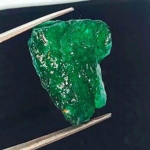 May include: A large, rough, green emerald gemstone. The emerald is held by tweezers against a black background.