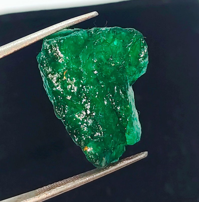 May include: A large, rough, green emerald gemstone. The emerald is held by tweezers against a black background.
