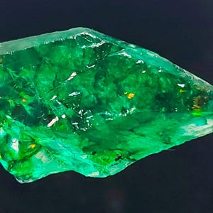 May include: A large, rough, green emerald gemstone with a slightly pointed tip. The emerald has a translucent, glassy appearance with visible inclusions.