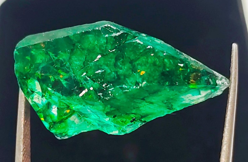 May include: A large, rough, green emerald gemstone with a slightly pointed tip. The emerald has a translucent, glassy appearance with visible inclusions.