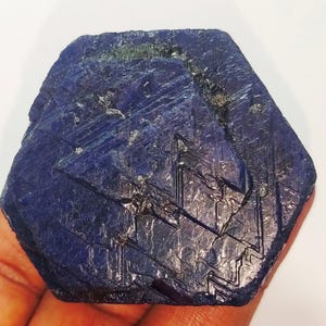 Puede incluir: Un gran cristal de zafiro azul oscuro con forma hexagonal facetada. El cristal tiene una textura rugosa y natural y muestra estrías intrincadas.