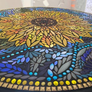 May include: A circular mosaic tabletop featuring a large central sunflower with yellow and orange petals. The surrounding pattern consists of various shades of blue, teal, and green glass tiles, with a border of small yellow and metallic gold tiles.