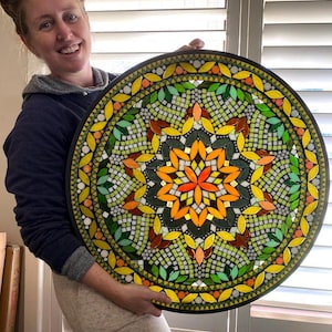 May include: A large circular mosaic wall art piece featuring a complex mandala pattern. The design incorporates vibrant shades of yellow, orange, and various tones of green glass tiles arranged in geometric shapes, including petals and squares.