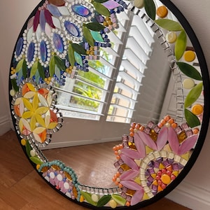 May include: A round mirror with a black frame featuring a decorative mosaic border. The mosaic consists of colorful glass pieces arranged in floral and leaf patterns, including shades of pink, yellow, blue, green, and orange tiles.