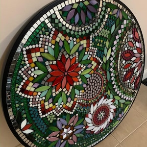 Könnte beinhalten: Ein kreisförmiges Mosaik-Wandkunstwerk mit einem komplexen Blumenmuster aus kleinen Glasfliesen in den Farben Rot, Grün, Violett und Weiß. Das Design ist von einem massiven schwarzen kreisförmigen Rahmen mit einer Umrandung aus weißen Fliesen umgeben.