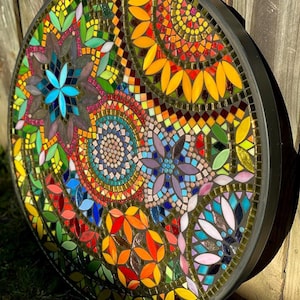 May include: A circular mosaic wall art piece featuring a vibrant array of multi-colored glass tiles arranged in intricate floral and geometric patterns. The design includes shades of yellow, orange, blue, green, and red, all encased in a black frame.