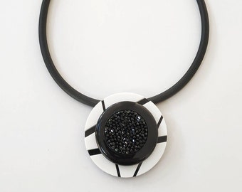 May include: A black and white geometric pendant necklace. The pendant features a circular design with a black center filled with small black beads. The necklace has a black rubber cord with a gold-colored clasp.