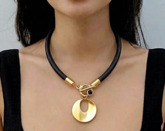 Puede incluir: Un collar de cordón negro con un cierre de palanca dorado y un colgante circular dorado. El collar se lleva alrededor del cuello, mostrando un cordón oscuro y liso que se conecta a un dije circular de tono dorado metálico en el centro.