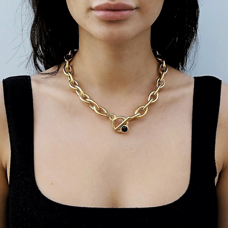 May include: A gold chain necklace with a large, chunky link design. The necklace has a toggle clasp with a black stone accent.