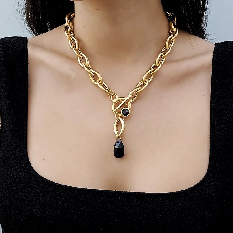 May include: A gold-coloured chain necklace with a toggle clasp and a black teardrop pendant. The necklace has a thick, linked chain design. The pendant is a black, teardrop-shaped stone.