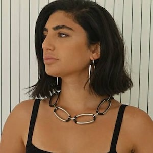 Puede incluir: Una mujer con el pelo negro corto lleva un collar de cadena plateado y negro. El collar tiene eslabones ovalados con un acento negro en cada eslab&oacute;n.