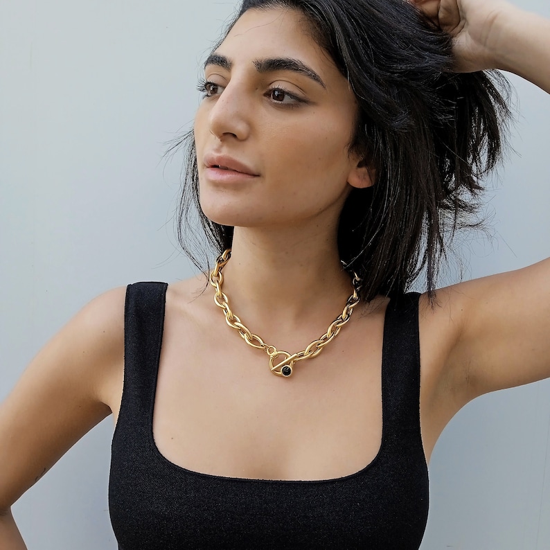 May include: A gold chain necklace with a large, chunky link design. The necklace has a toggle clasp with a black stone accent.