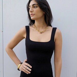 May include: A woman wearing a black sleeveless top with a gold chain necklace featuring a black stone. She is wearing a gold bangle bracelet on her wrist.