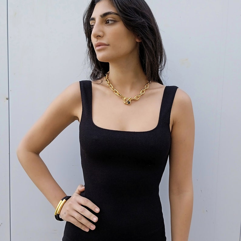 May include: A woman wearing a black sleeveless top with a gold chain necklace featuring a black stone. She is wearing a gold bangle bracelet on her wrist.