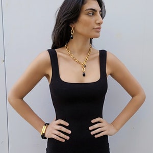 May include: A gold-coloured jewellery set comprising a necklace, earrings, and bracelet. The necklace features a chain design with a black teardrop pendant. The earrings and bracelet also have black accents. The woman is wearing a black dress.