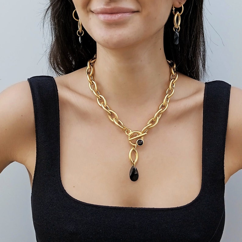 May include: Gold-coloured chain necklace with a toggle clasp and a black teardrop pendant. Matching gold earrings with black accents complete the jewellery set. The necklace features a chunky link design.