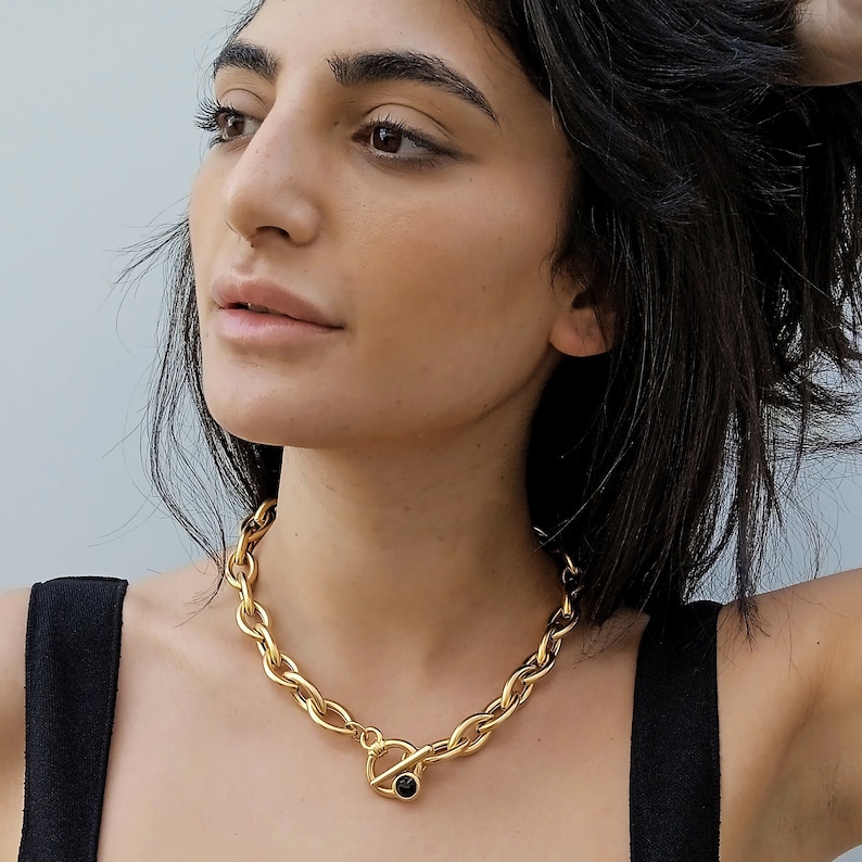 May include: A gold chain necklace with a large, chunky link design. The necklace has a toggle clasp with a black stone accent.