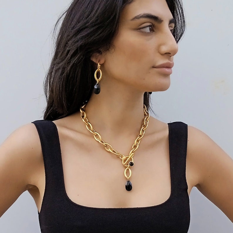 May include: A gold-coloured necklace and matching earrings set. The necklace has a chunky chain with a toggle clasp and a pendant with a black teardrop-shaped stone. The earrings have a similar design, with a black stone pendant.