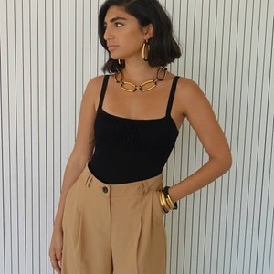 Puede incluir: Una mujer con un top negro de tirantes, un collar de cadena dorada y pendientes de aro dorados. Tambi&eacute;n lleva pantalones marrones y pulseras doradas.