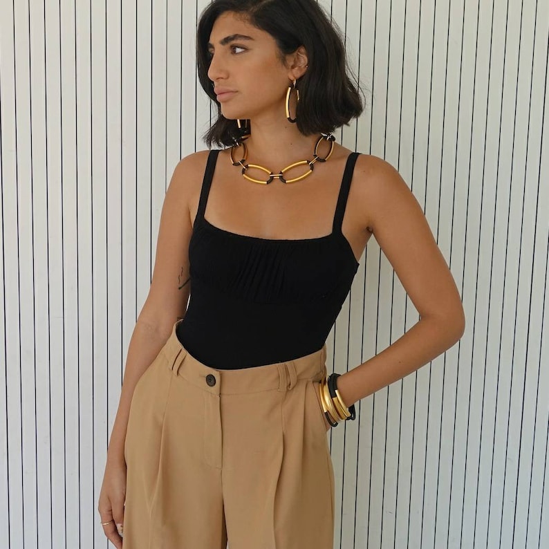 Puede incluir: Una mujer con un top negro de tirantes, un collar de cadena dorada y pendientes de aro dorados. Tambi&eacute;n lleva pantalones marrones y pulseras doradas.