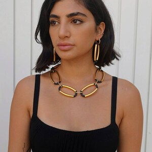 Puede incluir: Un collar de cadena dorada con detalles negros. El collar est&aacute; hecho de eslabones ovalados. La modelo lleva el collar con un top negro de tirantes.