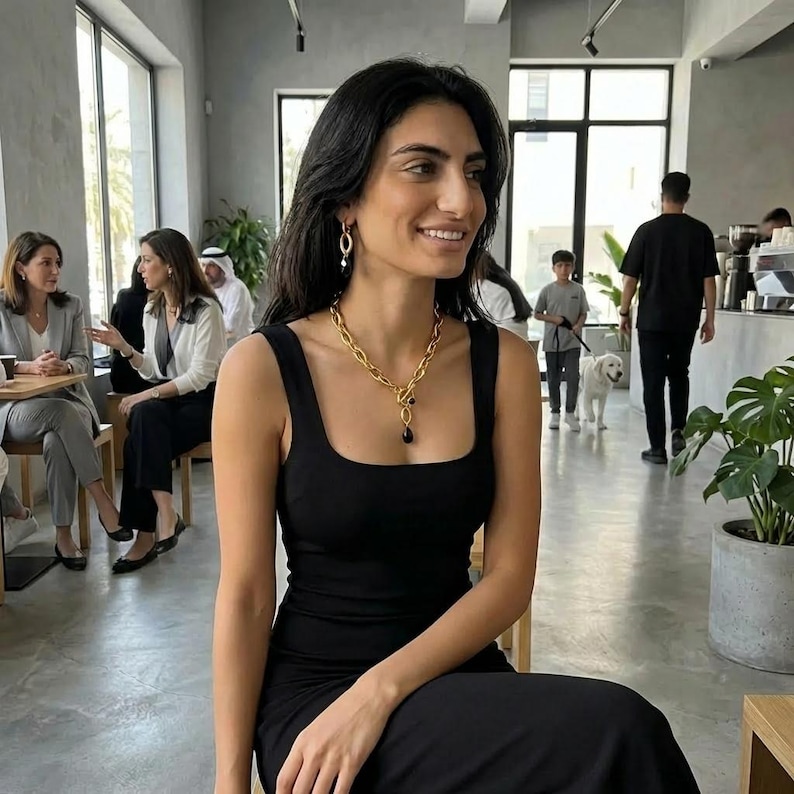May include: A woman in a black dress, adorned with a gold chain necklace featuring a black pendant and matching earrings. The necklace has a bold, chunky link design. The woman is smiling in a cafe.