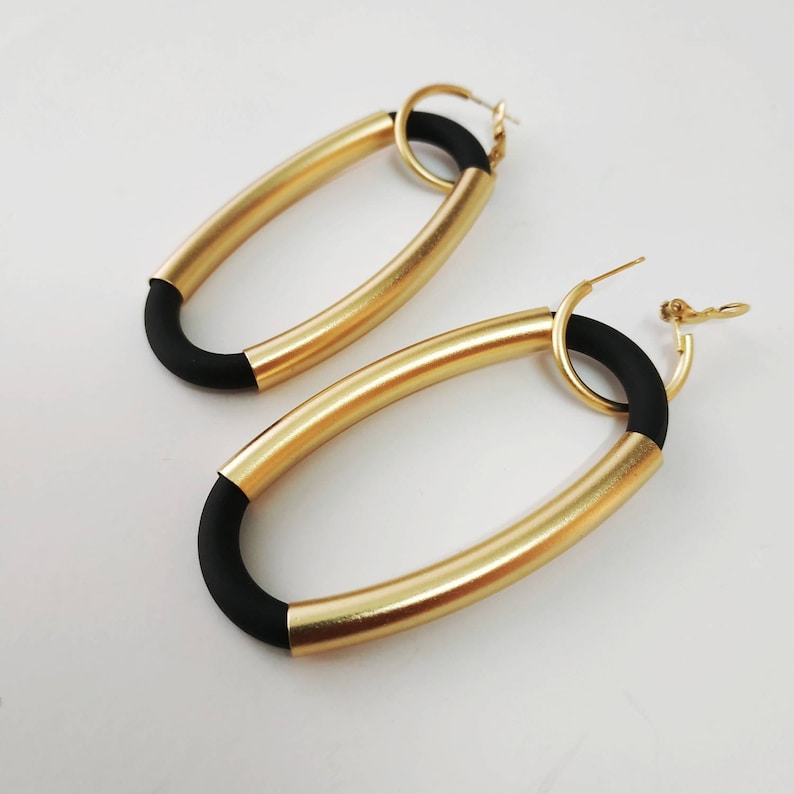 Puede incluir: Un par de pendientes de aro ovalados de oro con detalles de goma negra. Los pendientes tienen un dise&ntilde;o simple y moderno.