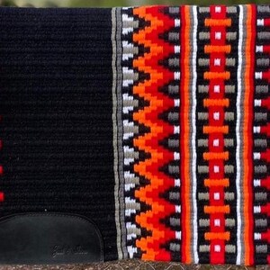 Op de afbeelding: Zwarte zadelpad met een geometrisch patroon in rood, oranje, wit en grijs. De zadelpad heeft een zwart lederen patch met de tekst "Trail Rider" erop.