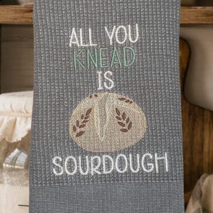 May include: A grey waffle-weave kitchen towel featuring white and sage green embroidered text that reads ALL YOU KNEAD IS SOURDOUGH, accompanied by a central embroidered graphic of a round loaf of bread with wheat stalk details.