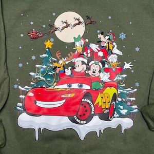 May include: Green long-sleeved sweatshirt with a Christmas-themed graphic featuring Mickey Mouse, Donald Duck, Goofy, and Pluto riding in a red Lightning McQueen car. The characters are wearing Santa hats and the car is driving through a snowy scene with a Christmas tree.