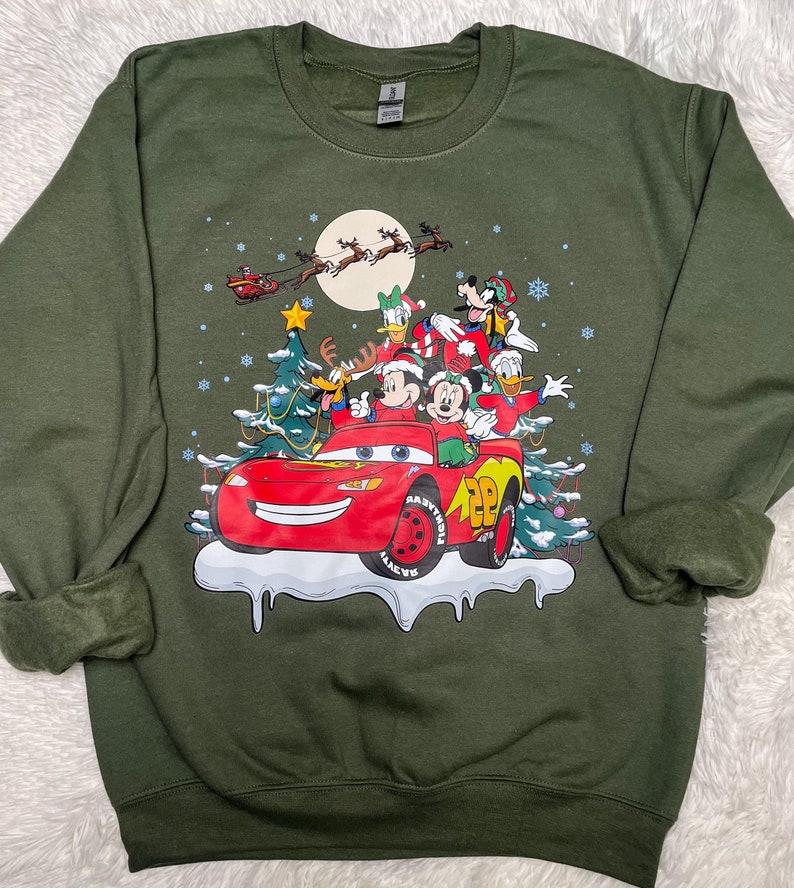 May include: Green long-sleeved sweatshirt with a Christmas-themed graphic featuring Mickey Mouse, Donald Duck, Goofy, and Pluto riding in a red Lightning McQueen car. The characters are wearing Santa hats and the car is driving through a snowy scene with a Christmas tree.