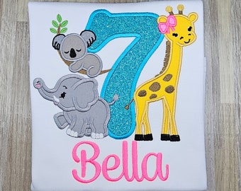 May include: A white t-shirt with a blue glittery number 7, a yellow giraffe with a pink bow, a gray elephant, and a gray koala. The shirt also has the name "Bella" in pink letters.