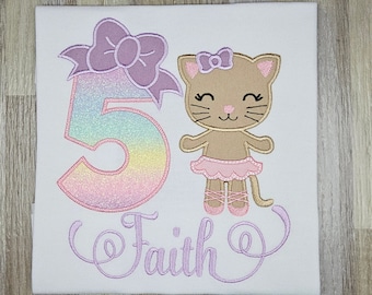 May include: A white t-shirt with a rainbow glitter number 5, a pink bow, and a brown ballerina cat with a pink bow. The name "Faith" is embroidered in purple below the design.