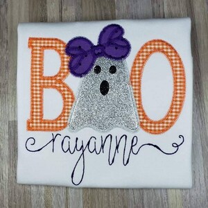 May include: White t-shirt with an orange and white gingham "BOO" with a silver glitter ghost wearing a purple bow. The name "rayanne" is embroidered below the ghost.