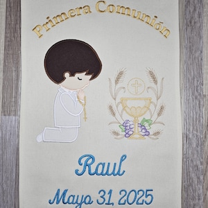 May include: A beige fabric banner with embroidered text that reads "Primera Comunión" in gold thread. The banner also features an embroidered image of a boy kneeling in prayer, a chalice, and grapes. The boy's hair is brown, his shirt is white, and his pants are gray. The text "Raul" is embroidered in blue thread below the image, and the date "Mayo 31, 2025" is embroidered in blue thread below the name.