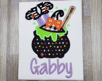 May include: A white t-shirt with an embroidered design of a black cauldron with green slime, a witch's hat with a black and white bow, and a broom. The text "Gabby" is embroidered in purple below the cauldron.