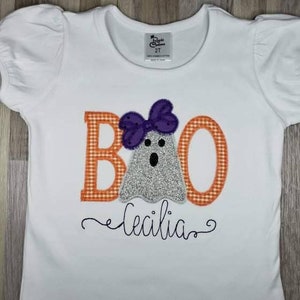 May include: White t-shirt with an orange and white checkered "BOO" with a ghost wearing a purple bow. The name "Cecilia" is embroidered below the ghost.