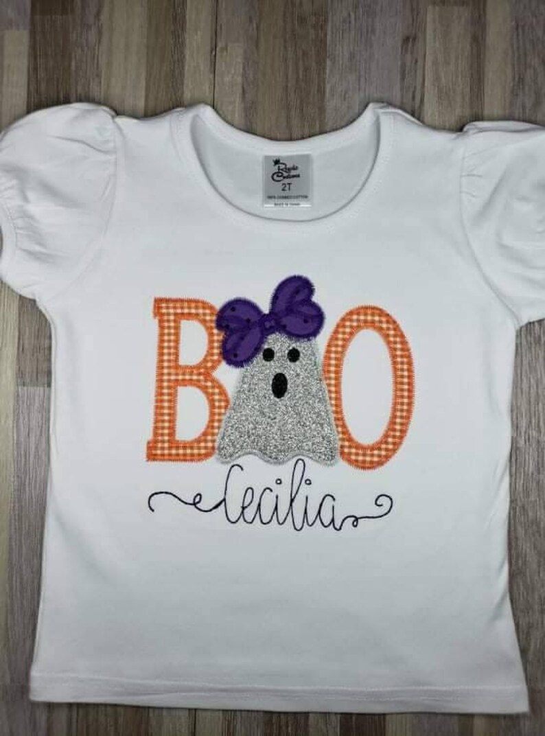 May include: White t-shirt with an orange and white checkered "BOO" with a ghost wearing a purple bow. The name "Cecilia" is embroidered below the ghost.
