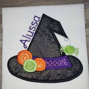 K&ouml;nnte beinhalten: Wei&szlig;es T-Shirt mit einem gestickten Hexenhut-Design. Der schwarze Hut hat eine glitzernde Textur, ein lila Band und orangefarbene und gr&uuml;ne Verzierungen. Der Name "Alyssa" ist lila gestickt.