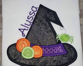 May include: White t-shirt with an embroidered witch hat design. The black hat has a sparkly texture, a purple band, and orange and green embellishments. The name "Alyssa" is embroidered in purple.