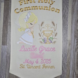 May include: A white banner with gold embroidery that says "First Holy Communion". A cartoon girl in a white dress is praying with a chalice and host in front of her. The banner also says "Lucille Grace", "May 4, 2025", and "St. Vincent Ferrer".