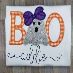 May include: White t-shirt with an embroidered design of the word "BOO" with a ghost wearing a purple bow. The name "addie" is embroidered below the design.